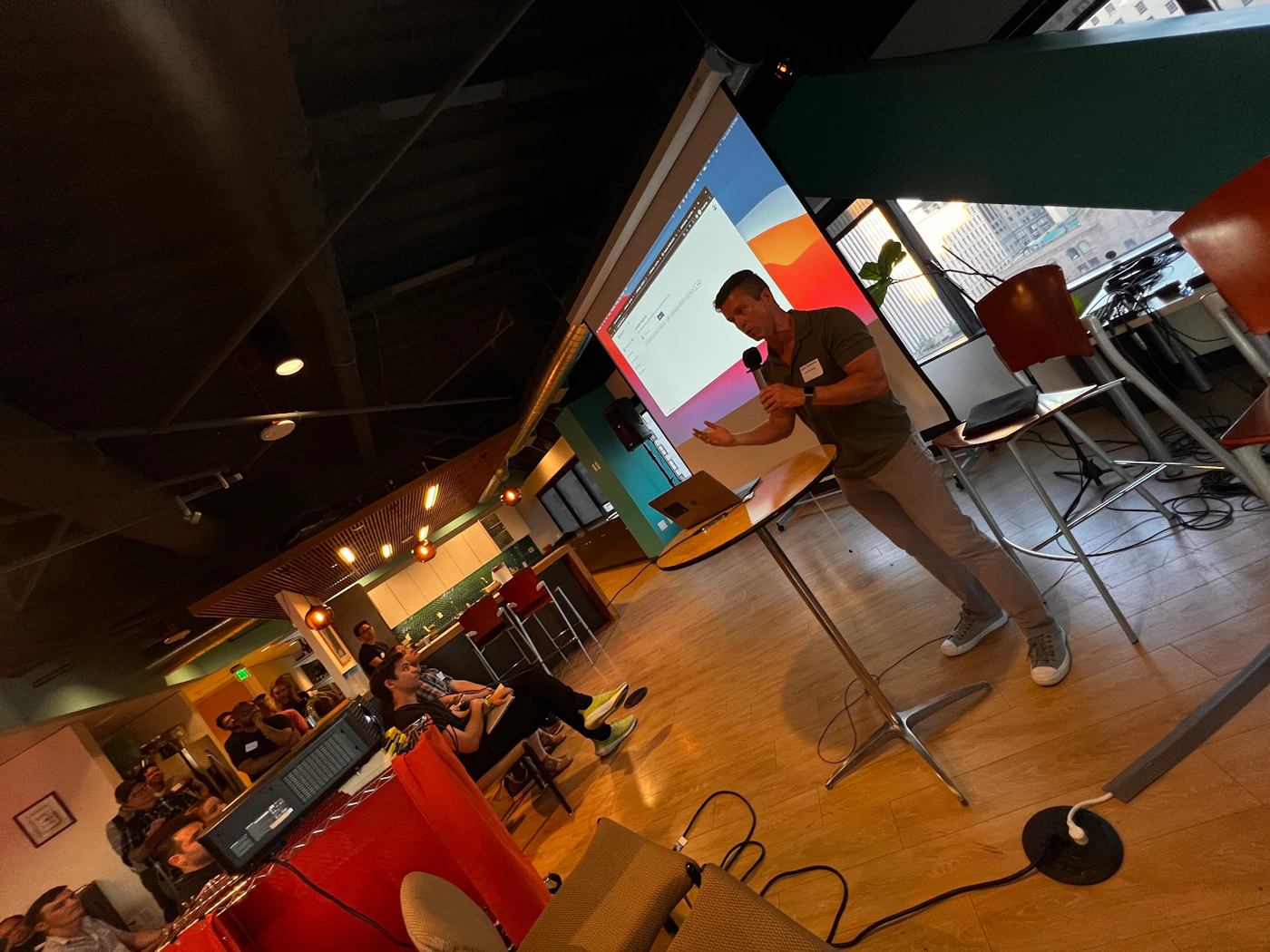 (Photo) A man is giving a presentation in a room with an audience. He is standing at a small table with a laptop and microphone, gesturing while speaking to the group. person | indoor | presentation, audience, projector | candid Note: This is a real-life captured image of a presentation setting, depicting people and their environment. It exhibits characteristics of a candid photograph.