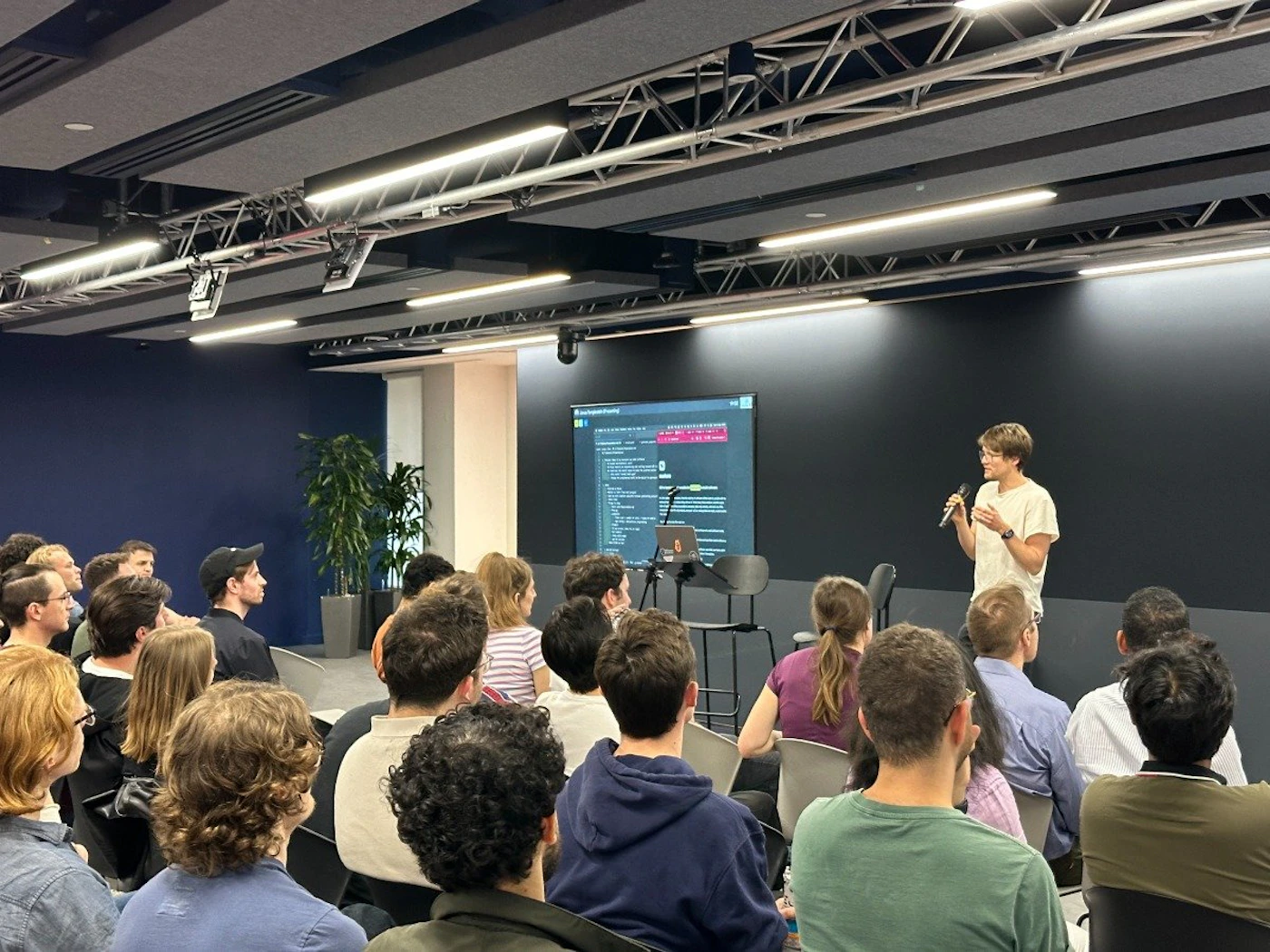 (Photo) A person stands on a small stage, holding a microphone and speaking to an audience seated in chairs.  A large screen displays what appears to be code or technical information. people | indoor | presentation, audience, screen | candid Note: The image is a real-life captured scene of a presentation, depicting people and a setting, which makes it a photograph.