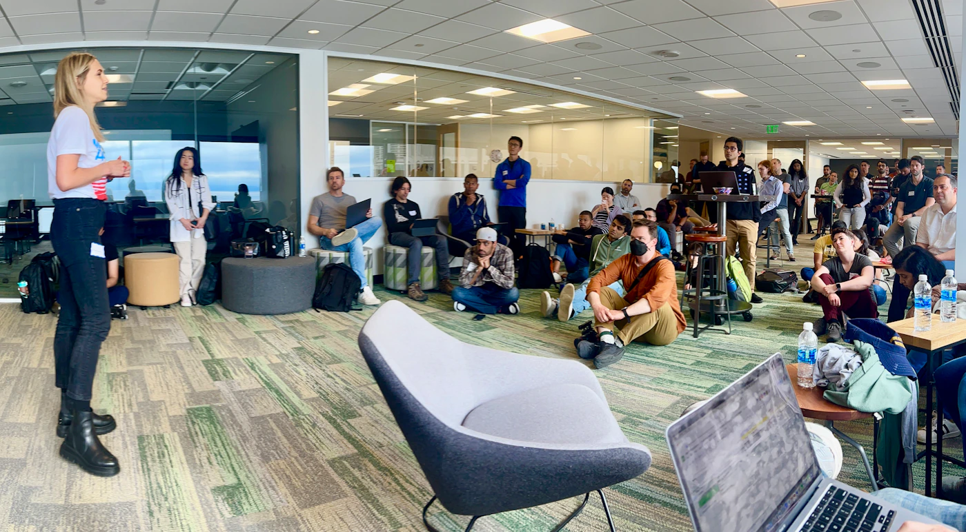 (Photo) A woman is presenting to a group of people in a modern office space. The audience is seated on chairs, stools, and the floor, listening attentively. people | indoor | presentation, office setting, audience | candid Note: The image is a real-life captured moment of people in an office setting, indicative of a photograph. It's not a drawn or digitally created image, nor a logo or banner.