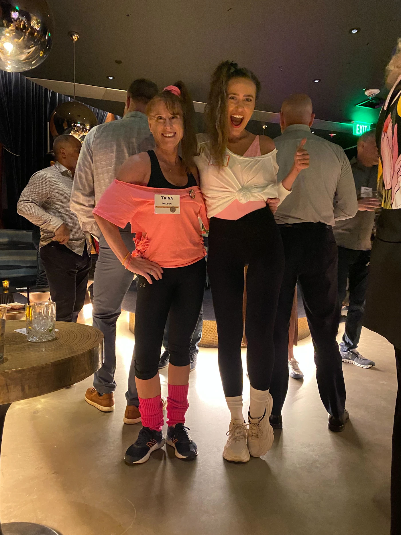(Photo) Two women dressed in 80s style workout attire pose for a photo in a dimly lit room with other people around them. Text: TRINA NELSON people | indoor | Two women in 80s attire, group of people, social gathering | candid Note: This is a photograph of people in a social setting.  It captures a moment in time with real people and a real environment.