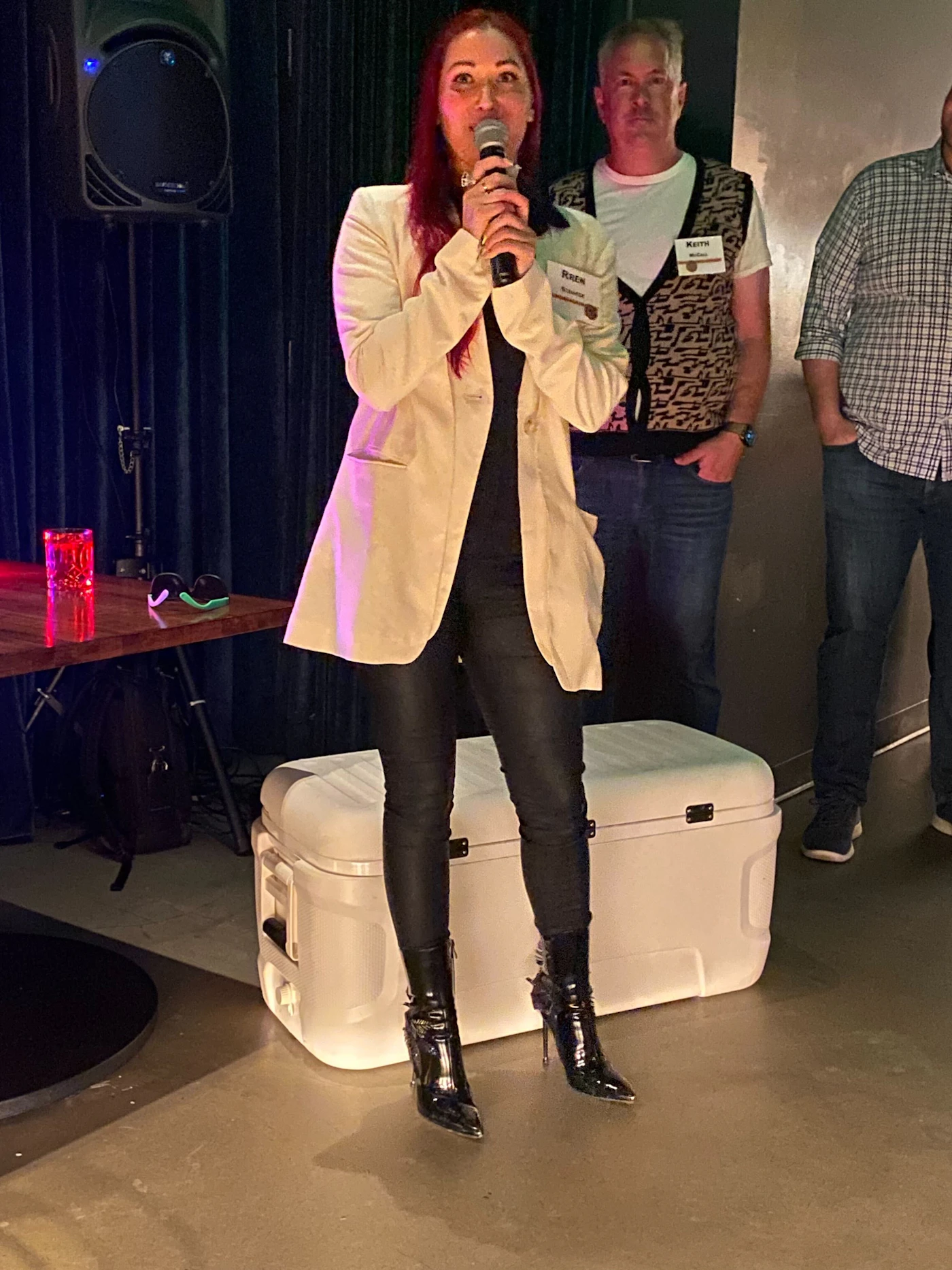 (Photo) A woman with red hair is speaking into a microphone on a stage, with two men standing behind her. Text: KEITH MAYER people | indoor | microphone, stage, audience | candid Note: This is a real-life captured image of people in an event setting, thus categorizing it as a photo.