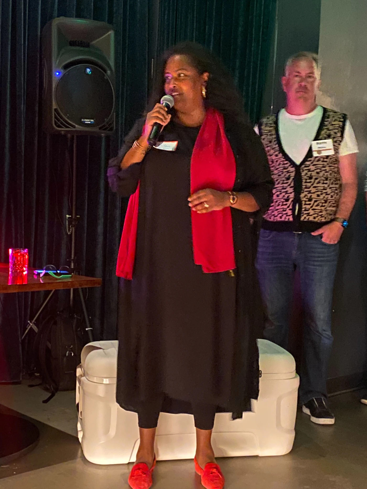 (Photo) A woman speaks into a microphone on a stage, with a man standing behind her. people | indoor | microphone, stage, audience | candid Note: The image is a real-life captured moment of people in an event setting, indicative of a photograph.