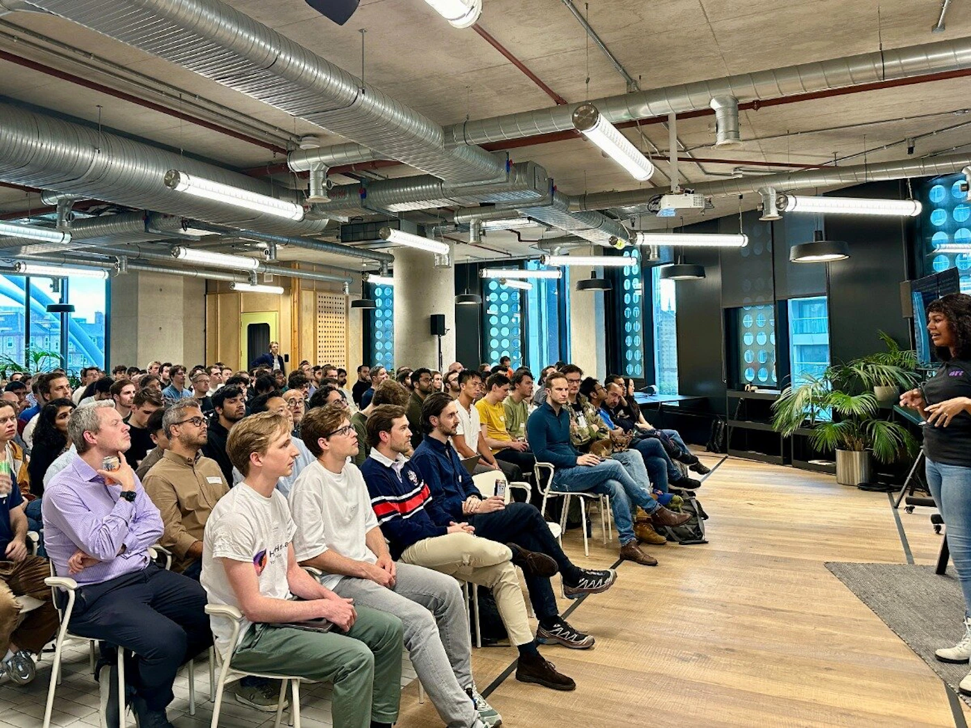 (Photo) A photo of a woman giving a presentation to a large audience in a modern office space. people | indoor | presentation, audience, office space | candid Note: The image is a real-life captured image of a scene, depicting people in an office setting, making it a photograph.