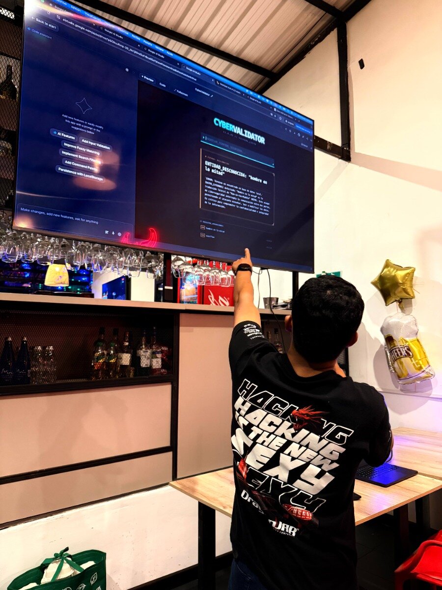 A person wearing a black t-shirt with a white graphic stands indoors, pointing toward a large, wall-mounted TV screen displaying a dark-themed application interface labeled 'CYBERVALIDATOR' in what appears to be a casual bar or meeting setting.. Text: Back to start Google AI Studio Code Followers CYBERVALIDATOR AI Features Add New feature or modify existing ones with a prompt. Try the suggestions below. Add Input Validation Improve Fuzzy Matching Implement Search Feature Add Command Prompt Persistence with LocalStorage Make changes, add new features, ask for anything ENTIDAD DESCONOCIDA: "Hombre en la edad" [Spanish text block regarding data and context] HACKING IS THE NEW SEXY Drag[on] [unreadable small text]