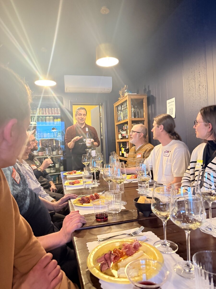 (Photo) A group of people are gathered around a long wooden table for a wine tasting event, listening to a speaker presenting a bottle of wine. Text: Hello Chris, Joel, Amy, THE MER..., Wine Club people at a wine tasting | indoor tasting room | wine glasses, food plates, speaker with wine bottle, name tags | candid | Colors: #2c3444, #d4af37, #5d4037, #f5f5dc Note: This is a candid photograph capturing a real-life social event in an indoor setting.