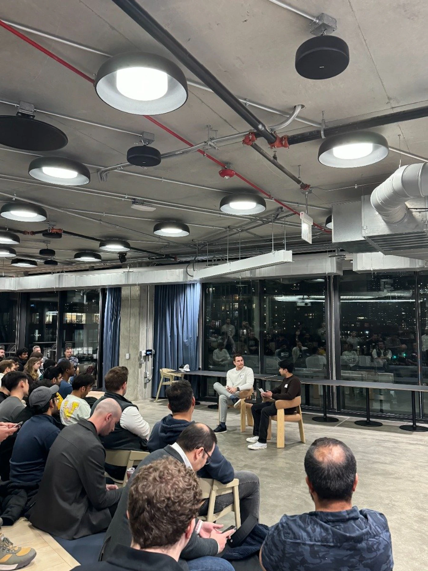 (Photo) A photo of a group of people sitting in an indoor space, seemingly listening to two men who are seated in chairs at the front. people | indoor | audience, speakers, presentation | candid Note: The image is a real-life captured scene of an event with people, indicating it's a photograph.  It's not a logo, illustration, banner, or handwriting.