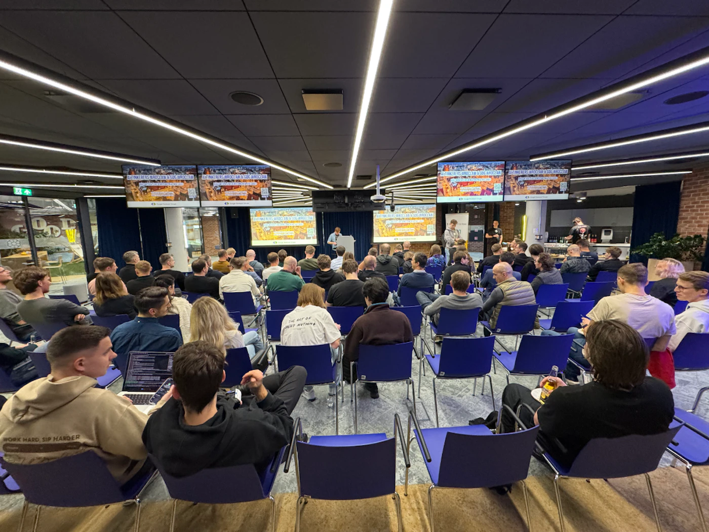 (Photo) A wide-angle photograph of a tech presentation or meetup event, showing a large audience seated in blue chairs facing a speaker at a podium and multiple display screens. Text: GDG Wroclaw & GDG Cloud Wroclaw & WTM Wroclaw, Google Developer Group, THE ART OF DOING NOTHING, WORK HARD, SIP HARDER, EXIT Tech community presentation or meetup | Indoor auditorium or large meeting room | Multiple display screens, rows of blue chairs, speaker at a podium, audience members with laptops. | Wide-angle candid shot | Colors: #0000FF, #2C2C2C, #FFFFFF, #8B8B8B Note: The image is a real-life photograph capturing a scene of people at an event.