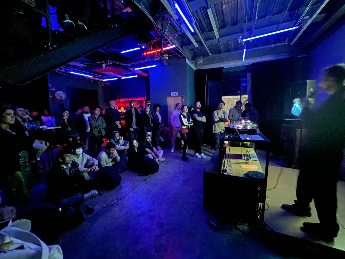 The image captures a dimly lit, intimate indoor event, likely a music or performance night, featuring a small audience gathered around a low stage. Attendees are sitting and standing, watching a figure standing near a DJ setup under intense blue and red neon lighting.