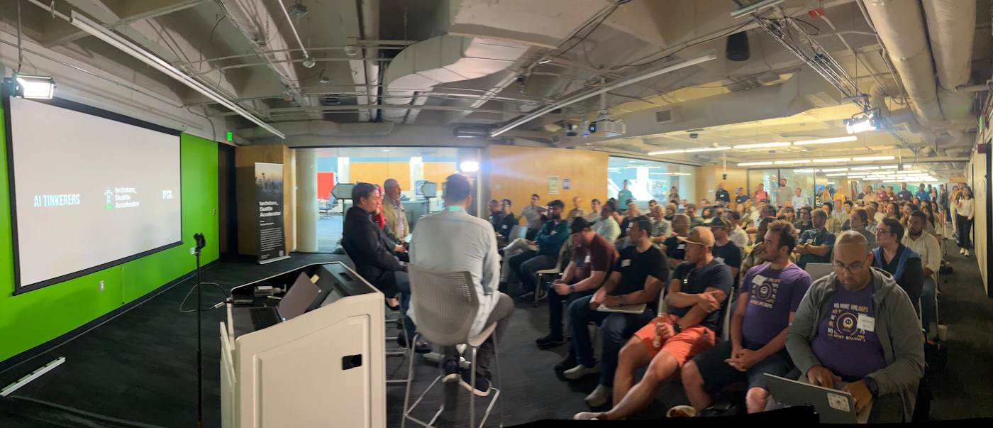 (Photo) A panoramic photo of a presentation or meeting in a modern office space.  A large audience is seated, facing a small panel of speakers on a stage area. Text: AI Tinkerer Industry, Built, Accelerator FGL people | indoor | presentation, audience, panel discussion | panoramic Note: The image is a real-life captured scene of an event with people, suggesting a photographic capture.