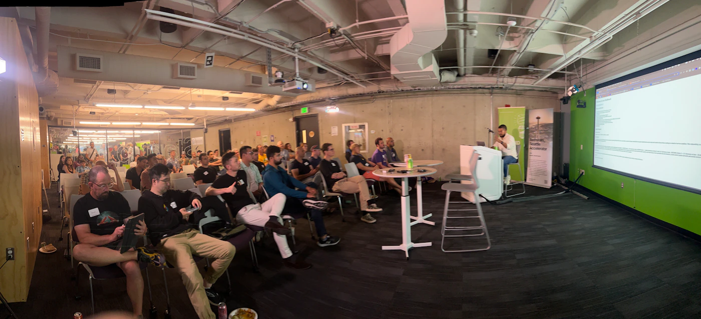 (Photo) A panoramic photo of a presentation or meeting in a large room.  An audience sits in chairs facing a speaker and a projected screen displaying code. people | indoor | presentation, audience, projector screen | panoramic Note: The image is a real-life captured scene of an event with people, making it a photograph.