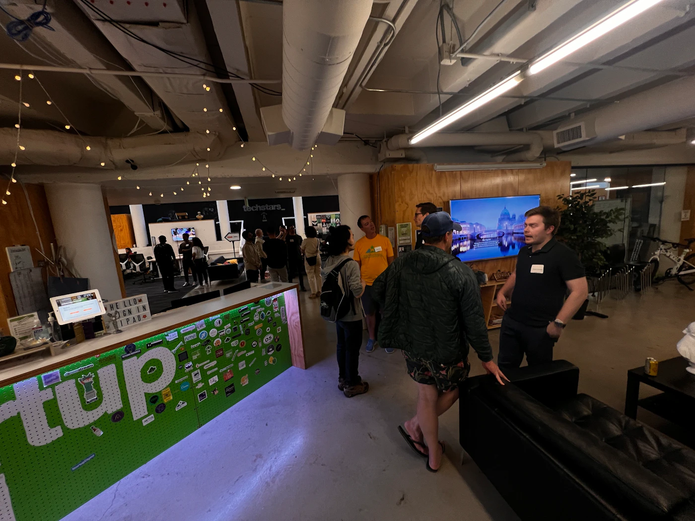 (Photo) A photo taken from a low angle inside a building, showing people gathered near a wall decorated with stickers and a large green sign. The image appears to be rotated. Text: up people | indoor | group of people, stickers on wall, large sign | candid Note: The image is a real-life captured scene of people in an indoor environment, making it a photograph.  It depicts a specific moment in time and is not a drawn or digitally created image.