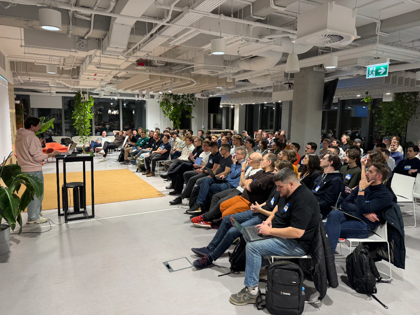 (Photo) A man in a pink hoodie delivers a presentation to a large audience seated in a modern, industrial-style office space. Text: ClickUp presentation | indoor office | large audience, industrial ceiling pipes, indoor plants, laptops | event photography | Colors: #D3D3D3, #2F2F2F, #F5E6E8, #4B6344 Note: This is a real-life photograph capturing a live event in an indoor setting with natural lighting and depth of field.