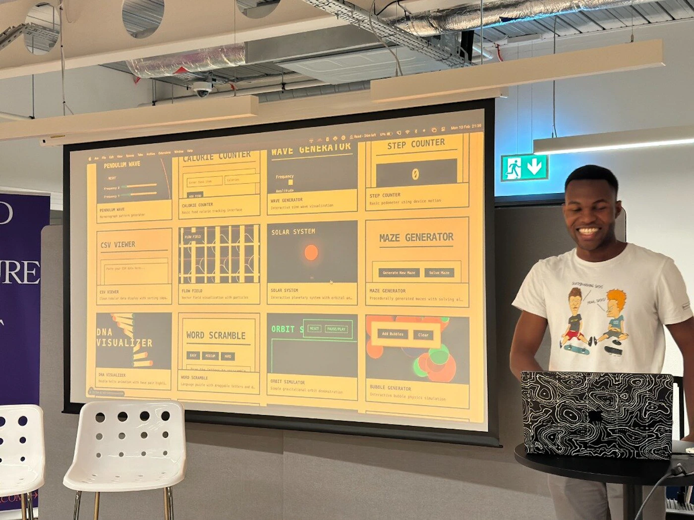 (Photo) A man stands next to a projected image showcasing a collection of interactive programs, likely during a presentation or demonstration. Text: PENDULUM MAVE CALORIE COUNTER WAVE GENERATUR STEP COUNTER 0 PENDULUM WAVE CALORIE COUNTER WAVE GENERATOR STEP COUNTER CSV VIEWER FLOW FIELD SOLAR SYSTEM CSV VIEWER FLOW FIELD SOLAR SYSTEM DNA WORD SCRAMBLE ORBIT VISUALIZER WORD SCRAMBLE ORBIT SIMULATOR DNA VISUALIZER MAZE GENERATOR BOBBLE GENERATOR MAZE GENERATOR BOBBLE GENERATOR person | indoor | projected image, presentation, interactive programs | candid Note: The image is a photograph of a person presenting a screen with interactive programs. It captures a real-life scene.