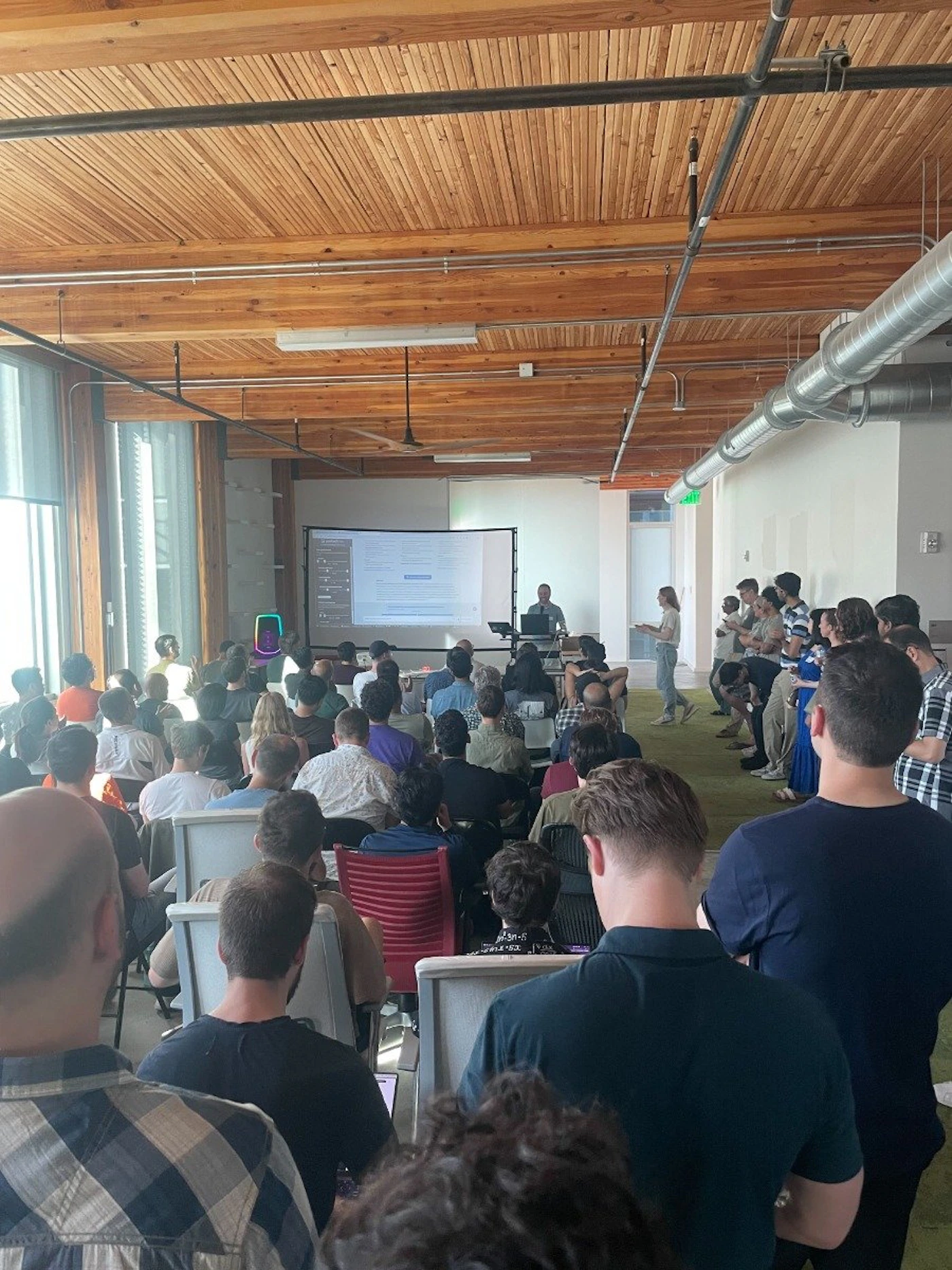 (Photo) A photo of a presentation in a room with a wooden ceiling.  A speaker is presenting to an audience seated in chairs. people | indoor | presentation, audience, wooden ceiling | candid Note: This is a real-life captured image of a presentation, depicting people and a setting, thus categorizing it as a photograph.