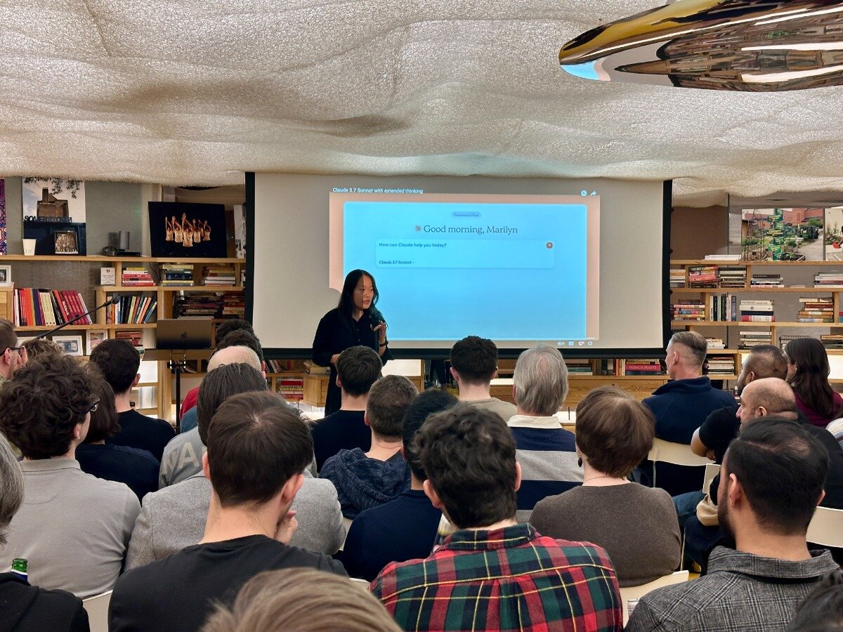 A woman presents to a seated audience in a room with bookshelves. A projected slide says "Good morning, Marilyn.". Text: Claude 3.7 format with extended thinking
* Good morning, Marilyn
How can Claude help?
on this topic?