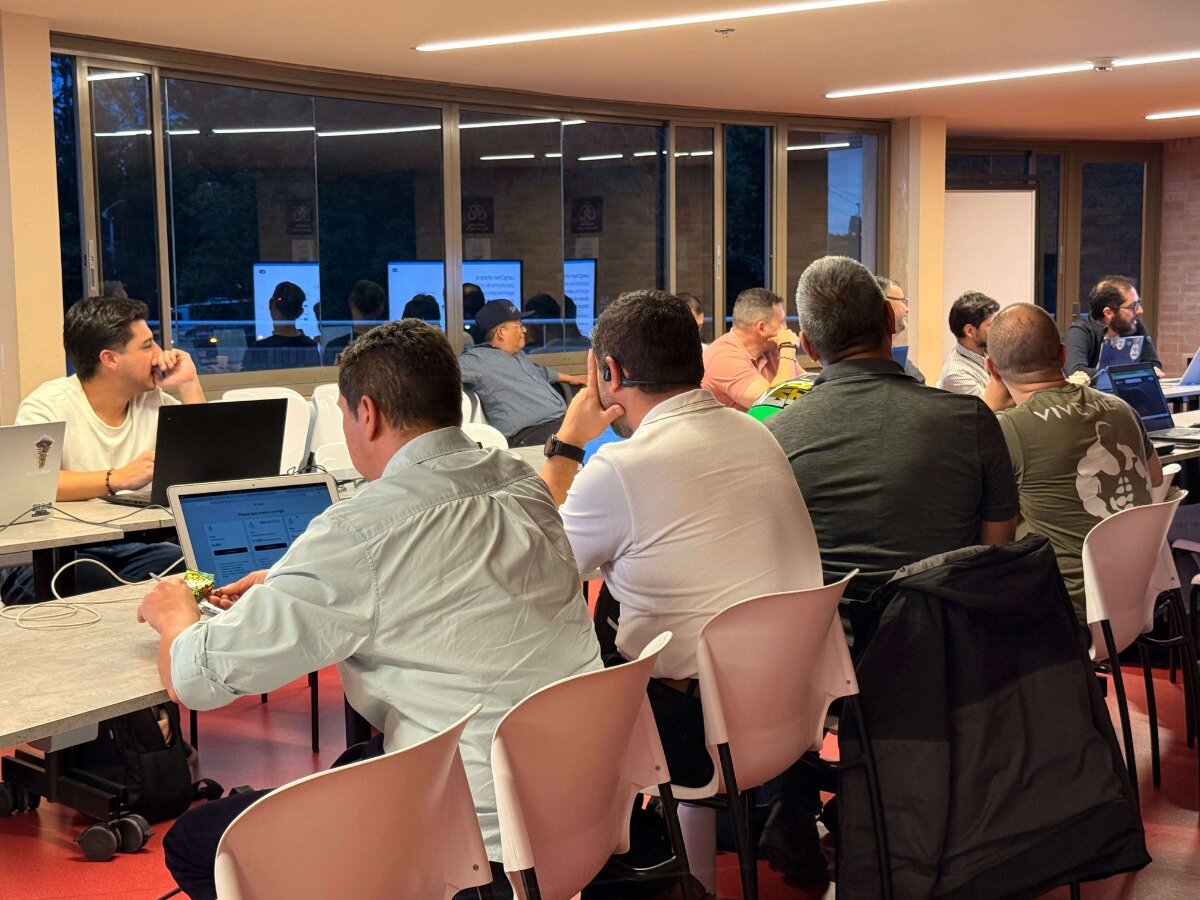 A group of people are seated at tables in a modern indoor setting, likely a workshop or office, working on laptops during the evening.. Text: VIVE VIVE