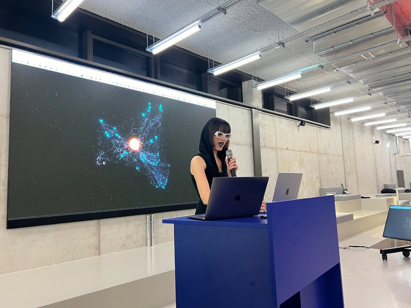 (Photo) A person with dark hair, wearing a black top and white sunglasses, is speaking into a microphone at a podium. A large screen behind them displays a graphic visualization, possibly of data or a network. person | indoor | presentation, data visualization | candid Note: The image is a real-life captured scene of a person presenting, making it a photograph. It's not a logo, illustration, or other graphic type.