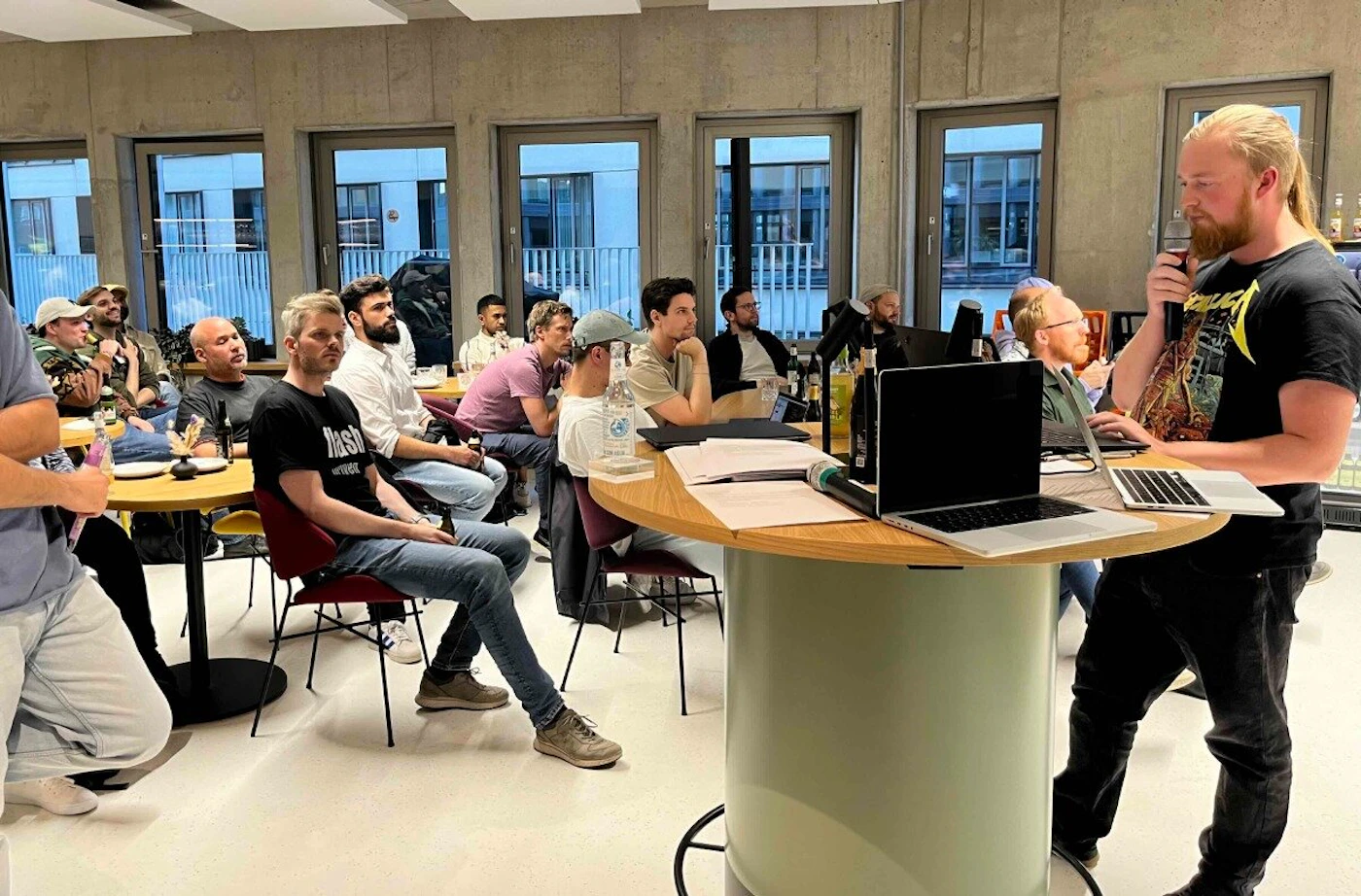 (Photo) A man is giving a presentation to a group of people in a casual office setting. The audience is seated at tables, listening and watching the presenter who is standing at a high table with laptops. people | indoor | presentation, group of people, office setting | candid Note: The image is a real-life captured moment of a presentation, showing people in a physical space interacting. This categorizes it as a photograph.