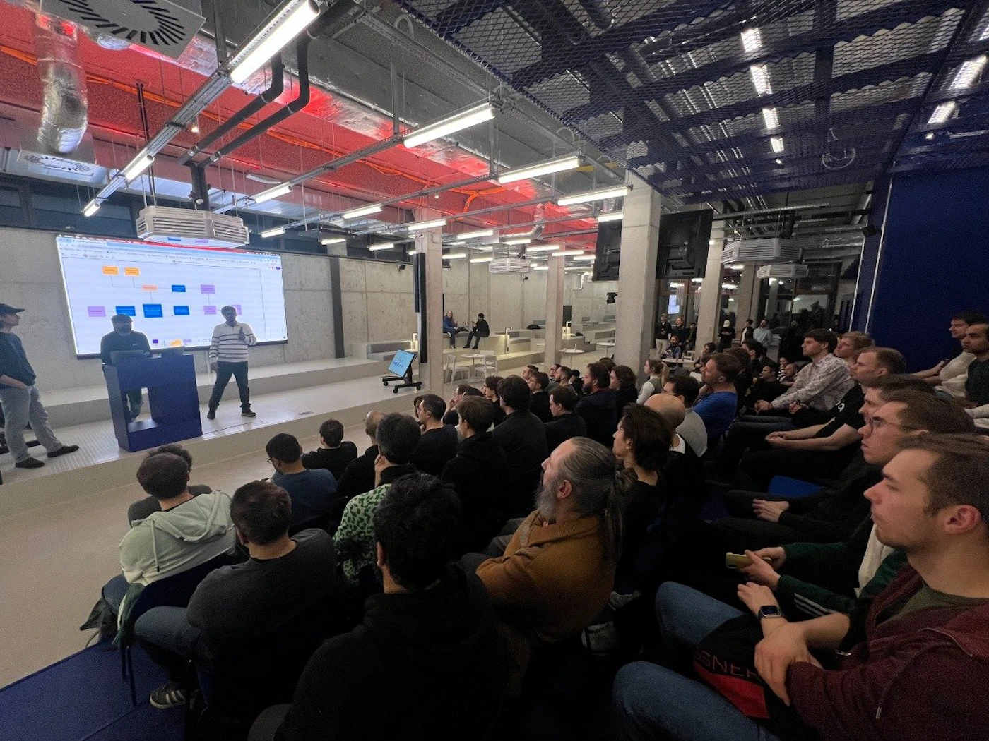 (Photo) A photo of a presentation taking place in a large, modern room.  An audience sits in rows of chairs facing a speaker on a small stage with a projected presentation behind them. people | indoor | presentation, audience, stage, projector screen | candid Note: The image is a real-life captured image of a scene, rather than a drawing, logo, or banner. Therefore, it is classified as a photo.