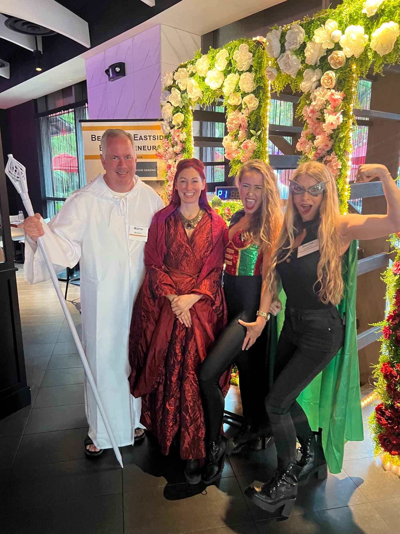 (Photo) Four people in costumes pose for a photo. They are standing in front of a floral archway. Text: EASTSIDE ENTREPRENEUR people | indoor | costumes, floral archway | candid Note: The image is a real-life captured moment of people in costumes, clearly a photograph and not a digitally created image.