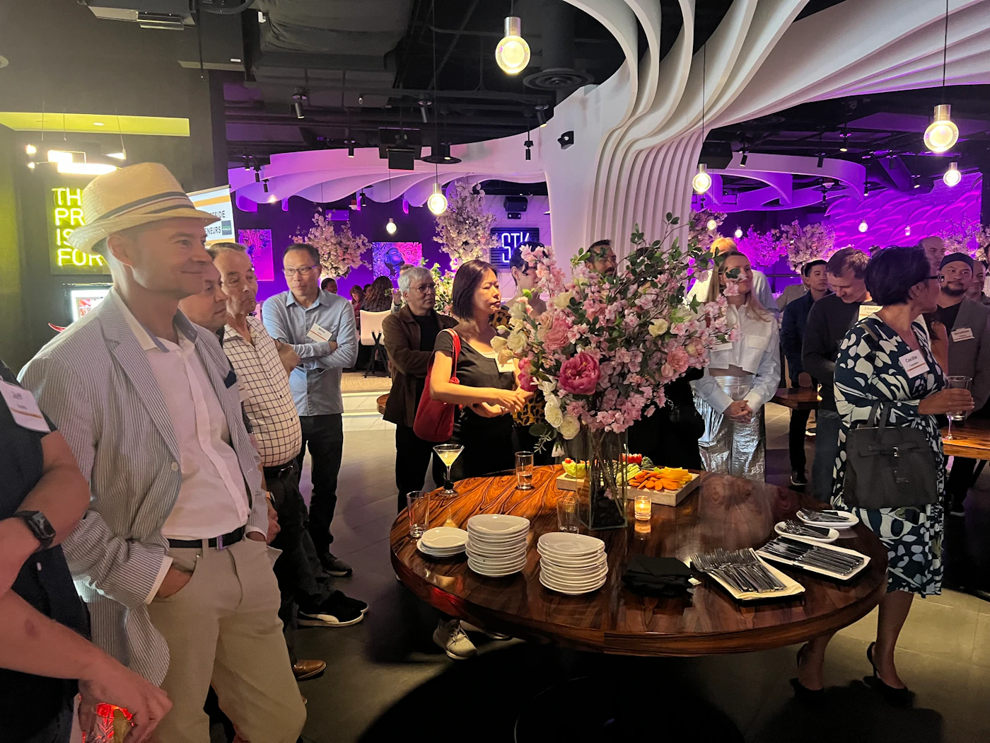 (Photo) A group of people are gathered in a dimly lit venue, possibly a restaurant or bar, socializing around a table with food and drinks. Text: TH PR IS FOR people | indoor | group of people, social gathering, food and drinks | candid Note: The image is a real-life captured scene of an event with people interacting in a physical space. It exhibits characteristics of a candid photograph.