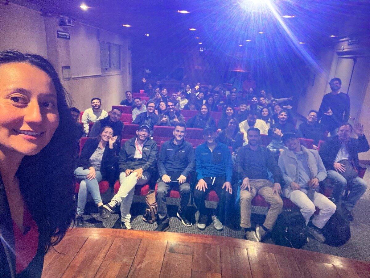 A group of people are gathered in an indoor auditorium or lecture hall, with a woman in the foreground taking a selfie that captures the audience behind her. A bright blue light beam from a projector illuminates the upper part of the room.