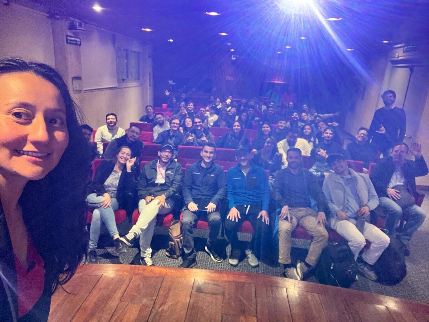 (Photo) A group of people are gathered in an indoor auditorium or lecture hall, with a woman in the foreground taking a selfie that captures the audience behind her. A bright blue light beam from a projector illuminates the upper part of the room. group of people, audience | indoor, auditorium/lecture hall | large group of diverse individuals, projector light beam, tiered seating, woman taking a selfie, many people smiling | candid, group photo, low light conditions | Colors: #4A4AFB, #8B1A1A, #8B4513, #F0C9B0, #2C2C2C Note: The image is a direct capture of a real-world scene featuring people and an environment, consistent with the definition of a photograph rather than an illustration, logo, banner, or handwriting.