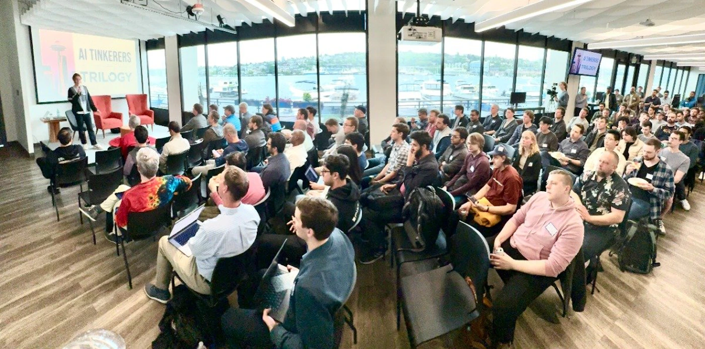 (Photo) A speaker presents to a large audience in a modern room with large windows overlooking a body of water. Text: AI Tinkerers TRILOGY people | indoor | presentation, audience, large windows, view of water | wide-angle Note: The image is a real-life captured scene of a presentation, depicting people, a venue, and projected text. This makes it a photograph.