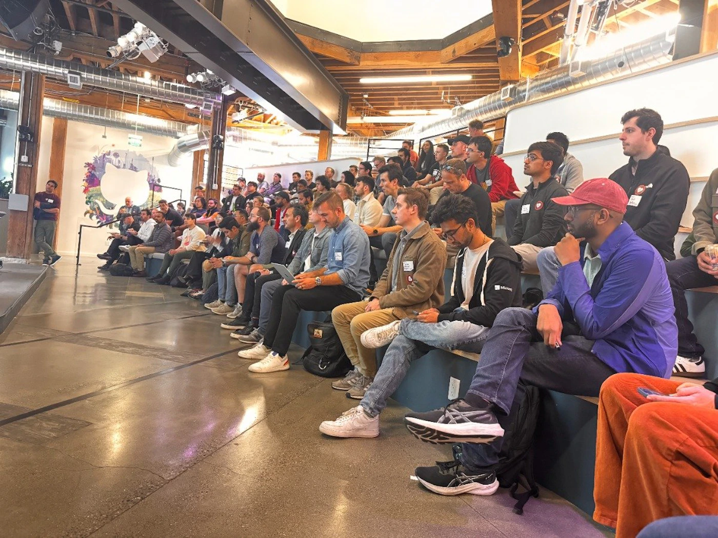 (Photo) A photo of a large group of people sitting in tiered seating, likely attending a presentation or meeting in a modern, industrial-style space. people | indoor | large group, tiered seating, presentation setting | candid Note: The image is a real-life captured scene of people in a specific setting, indicative of a photograph. It's not a drawing, logo, or banner.