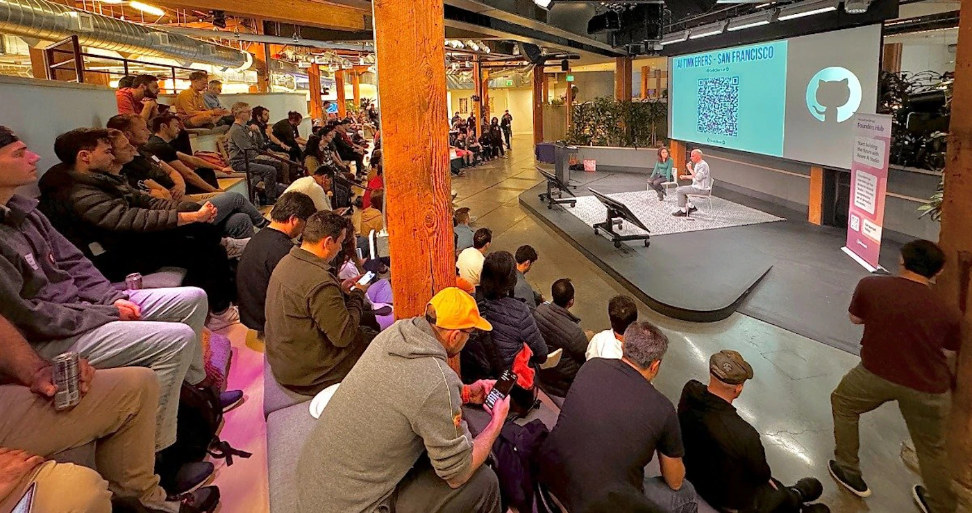 (Photo) A photo of an audience watching a presentation in a large, modern space. Two presenters are on a stage in front of a large screen. Text: AI FUNDRAISERS - SAN FRANCISCO people | indoor | presentation, audience, stage, screen | candid Note: The image is a real-life capture of an event, depicting people in a physical setting. This clearly identifies it as a photograph.