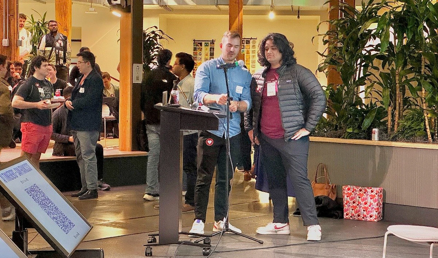 (Photo) Two individuals stand near a podium on a stage, seemingly addressing a small audience in a casual indoor setting. people | indoor | podium, audience, casual atmosphere | candid Note: The image captures a real-life scene with people, suggesting a photograph rather than a digitally created image or designed graphic.