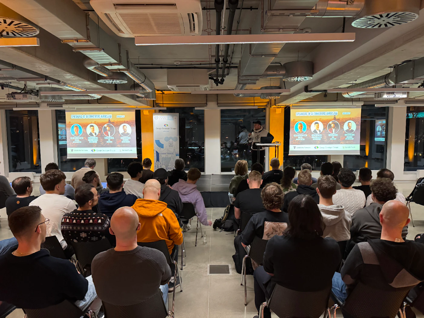 (Photo) A photograph of a tech meetup titled 'AI Tinkerers Wrocław' showing an audience seated in a modern industrial-style room watching a presentation. Text: SPEAKERS @ AI TINKERERS WROCŁAW KACPER SKONIECZNY MATEUSZ KRAJEWSKI PAWEŁ WYBIERAŁA ŁUKASZ KRAKOWIAK Google Developer Groups Code for the Future Daftcode Wrocław tech meetup/presentation | indoor modern office space | audience, projection screens, speaker, industrial ceiling | candid | Colors: #d3d3d3, #f9b233, #212121 Note: This is a real-life photograph capturing a scene of people attending an event in a physical space.