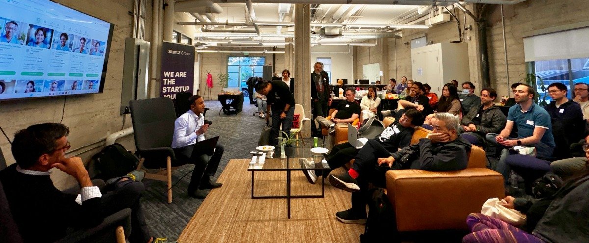 A group of people are gathered in a modern office space, some seated on couches and chairs, apparently listening to a presentation or discussion. A large screen displays a grid of headshots.. Text: Start12 Group
WE ARE
THE
STARTUP
YOU