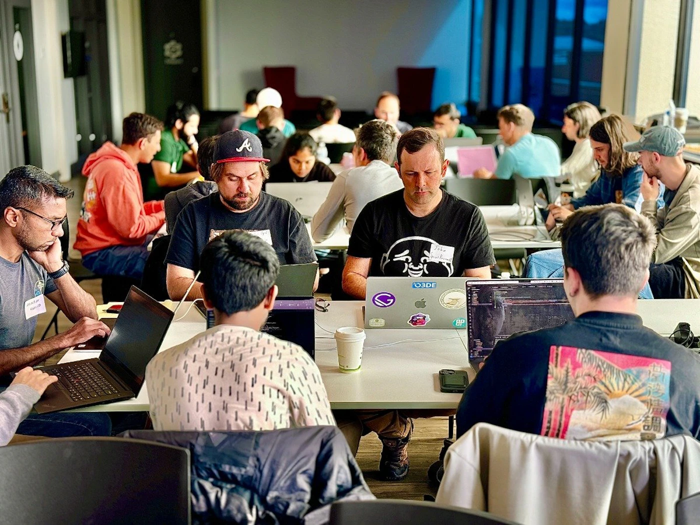 (Photo) A group of people are sitting at tables, working on laptops. They appear to be in a collaborative workspace or attending a workshop. people | indoor | multiple people, laptops, tables, collaborative workspace | candid Note: The image is a real-life captured scene of people in a workspace, making it a photograph. It's not a staged image or any other visual type.