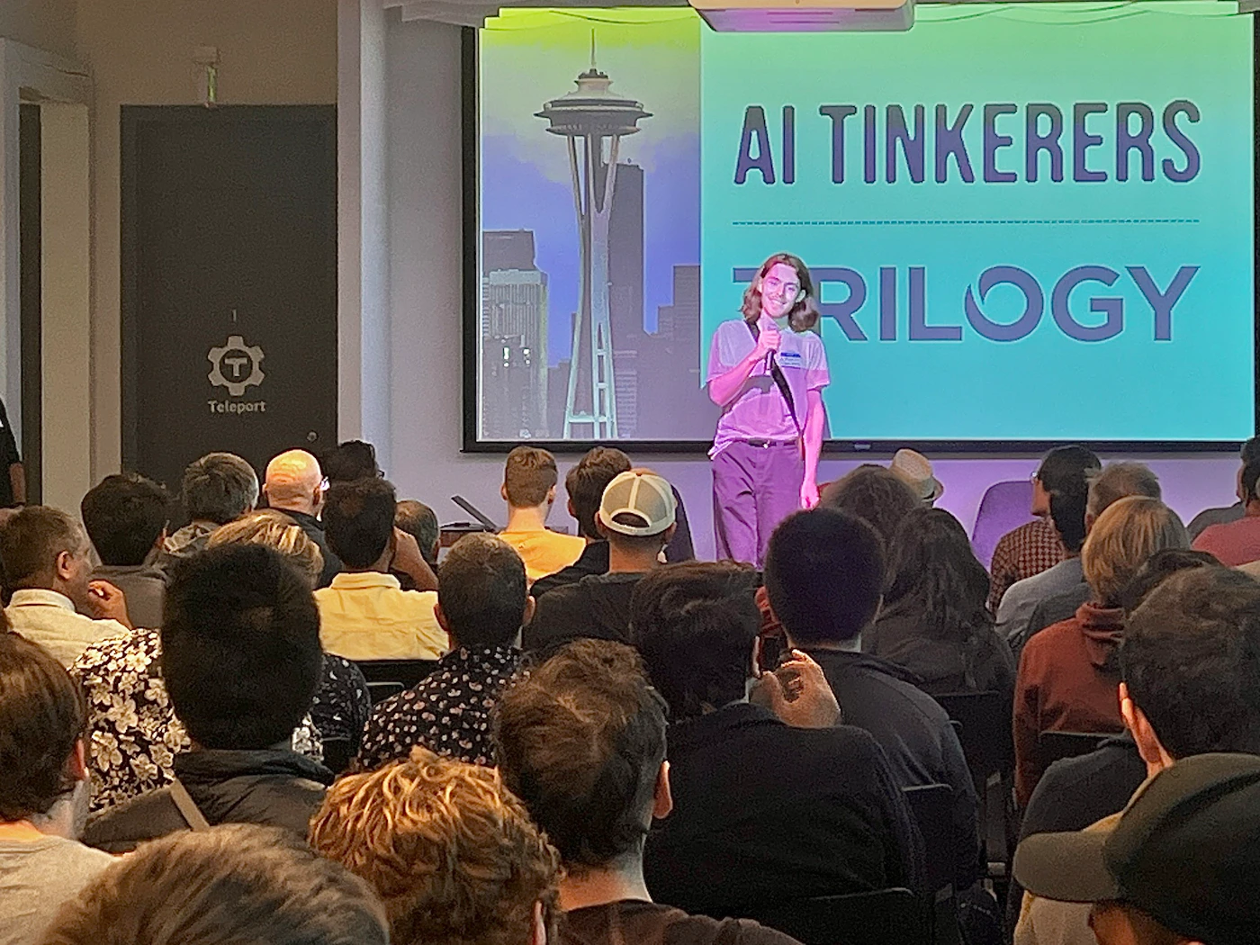 (Photo) A person stands on a stage in front of a projected screen, addressing an audience seated in rows of chairs. The screen displays the words "AI TINKERERS" and "TRILOGY" along with an image of the Space Needle. Text: AI TINKERERS TRILOGY Teleport people | indoor | presentation, audience, projected screen | candid Note: The image is a real-life captured moment of a presentation or event, depicting people and a setting, thus categorizing it as a photograph.