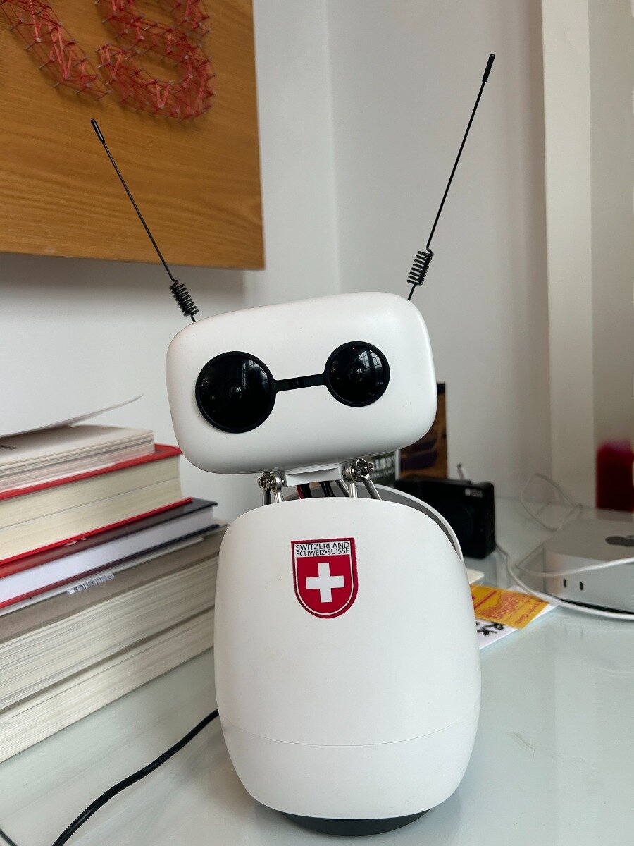 A small, white, rounded robot with two black circular eyes and thin wire antennas, sitting on a desk next to a stack of books.. Text: SWITZERLAND SCHWEIZ-SUISSE