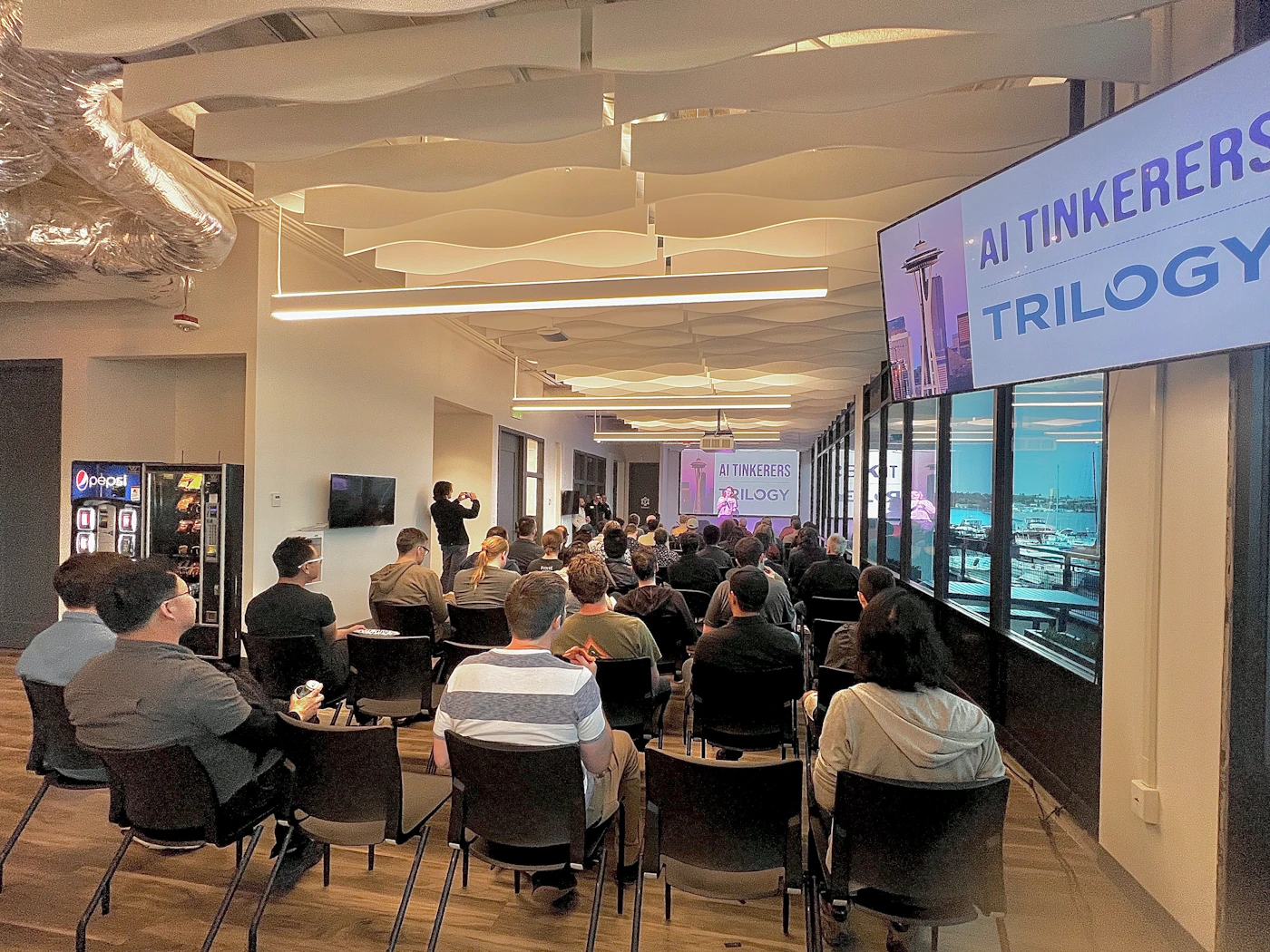 (Photo) A group of people are sitting in chairs in a modern office space, watching a presentation. A banner or screen displays "AI Tinkerer" and "Trilogy". Text: AI Tinkerers TRILOGY people | indoor | presentation, audience, office space | candid Note: The image is a real-life captured scene of an event or presentation, making it a photograph. It is not a logo, illustration or other created image type.
