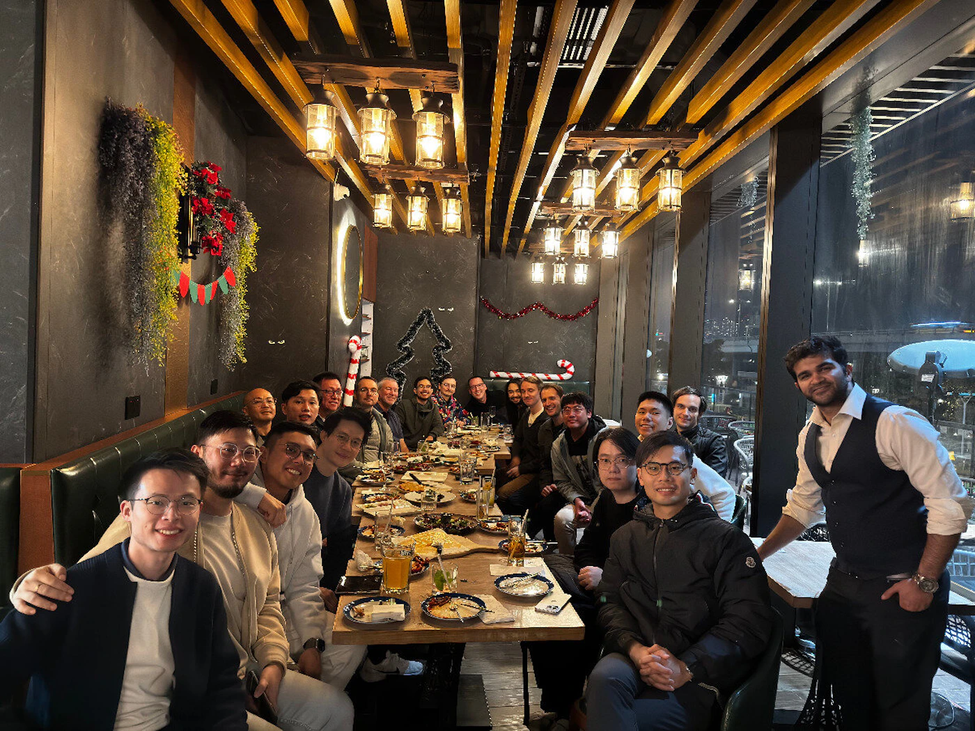 (Photo) A group of approximately twenty people, mostly men, are seated closely around a long table in a dimly lit restaurant or bar, appearing to be enjoying a social gathering or dinner. The venue features dark walls, wooden ceiling beams, lantern lighting, and subtle holiday decorations. A large group of people (social gathering/team dinner) | Indoor restaurant or bar, featuring dark, textured walls and large windows overlooking an outdoor area at night. | Low, warm lighting from hanging lanterns; holiday decorations (wreaths, candy canes, garlands); long communal table filled with food and drinks. | Group portrait/candid, taken in low light conditions. | Colors: #333333, #7F5539, #C8A040, #4B6A4B Note: The image is a captured moment of real people gathered in a physical setting, characteristic of a photograph.