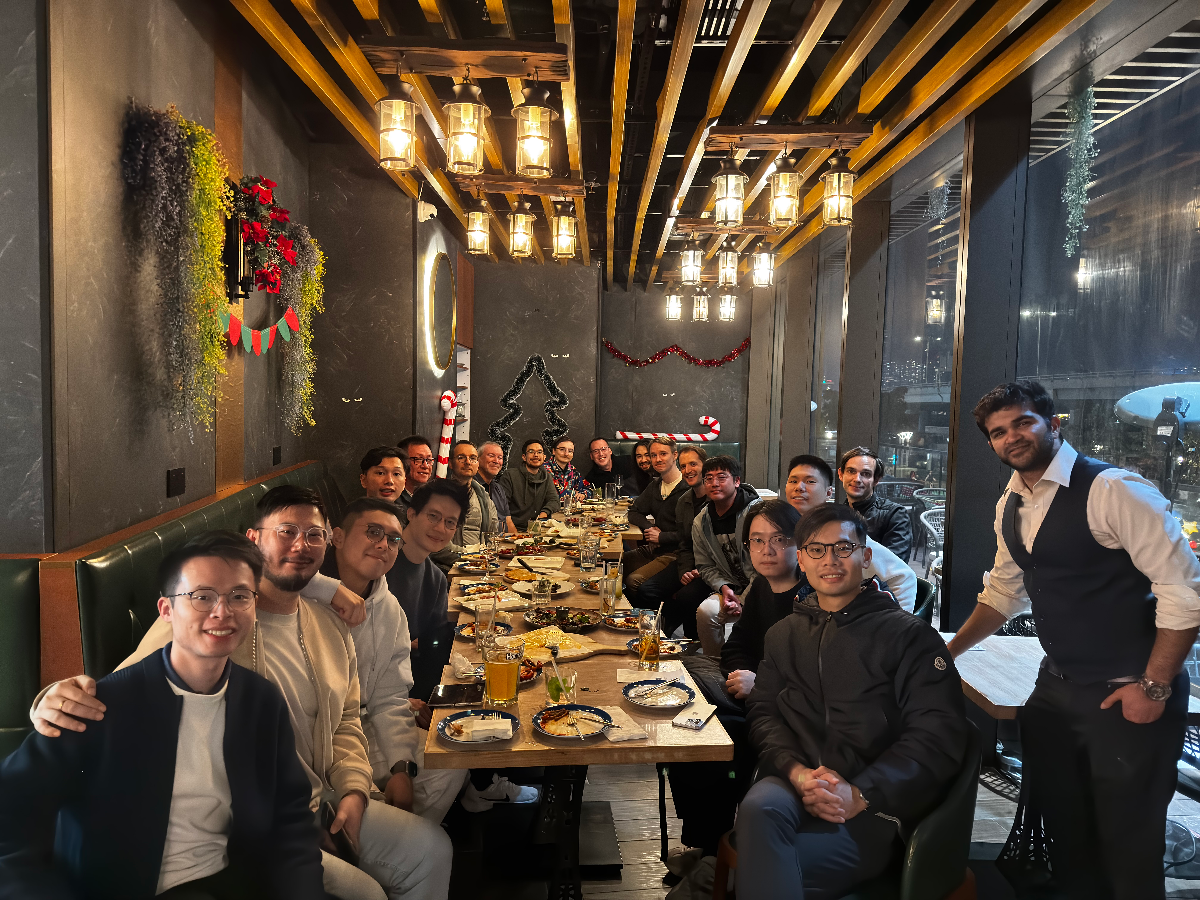 A large group of people is gathered around a long dining table in a warmly lit restaurant, which is decorated with holiday elements like wreaths and garlands.