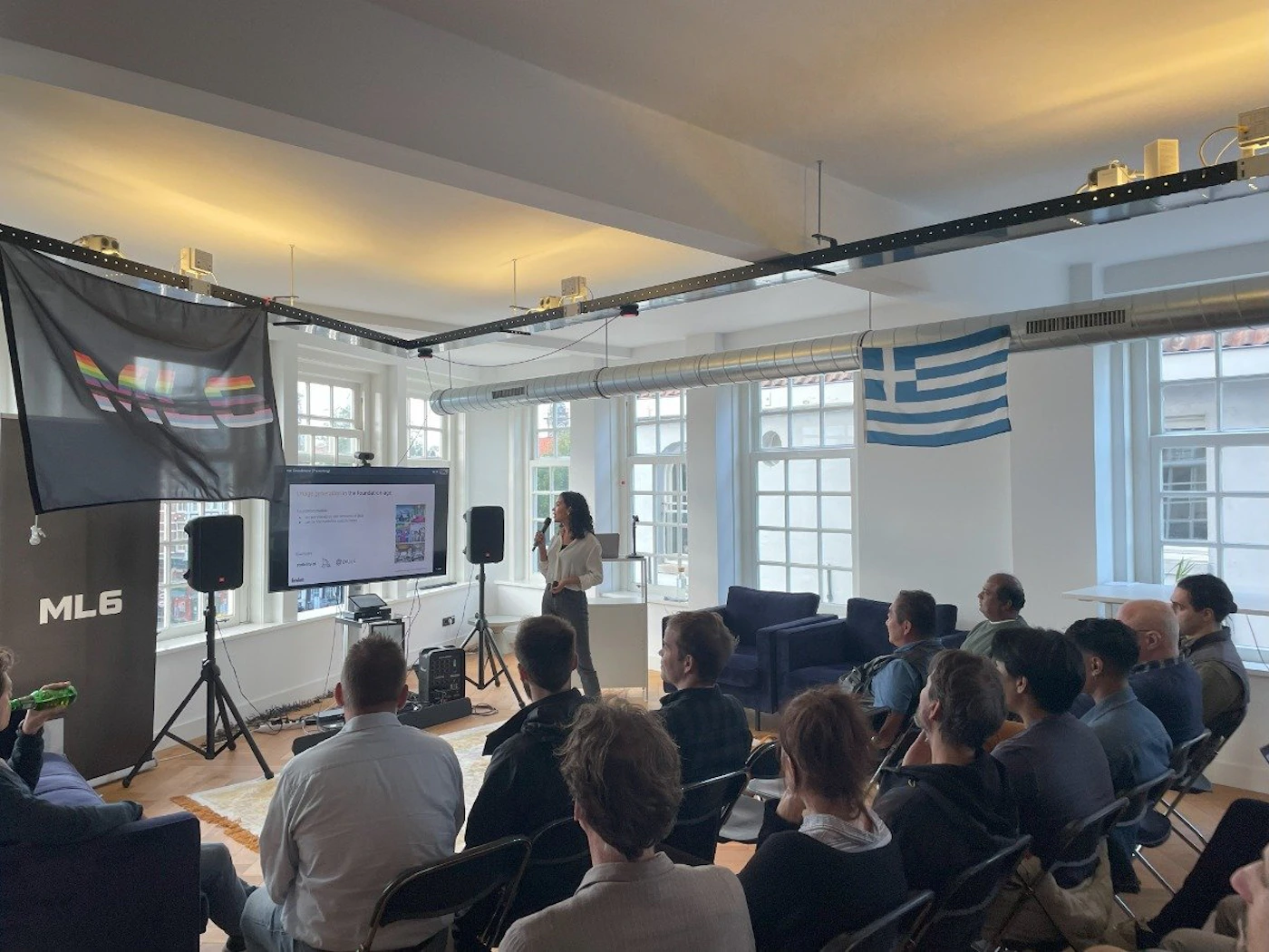 (Photo) A woman is presenting to a small audience in a room.  There are flags hanging from the ceiling, a screen displaying a presentation, and people sitting in chairs. Text: ML6 people | indoor | presentation, audience, flags | candid Note: This is a real-life captured image of a presentation setting, making it a photograph.  It's not a staged image or other visual media type.