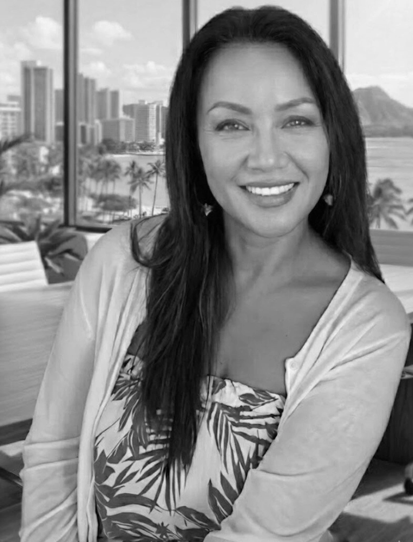 (Photo) A black and white portrait photograph of a smiling woman with long dark hair, wearing a cardigan over a patterned top, set against a window view of a tropical cityscape. person (adult female) | indoor (near a window) with an outdoor background (cityscape, ocean, and mountains) | The background appears to be a view of Waikiki/Diamond Head in Honolulu; the image is processed in black and white. | portrait (headshot/upper body), monochrome | Colors: #000000, #FFFFFF, #808080 Note: The image is a realistic capture of a person in a specific setting, using photographic techniques and rendered in monochrome, making it a photo.