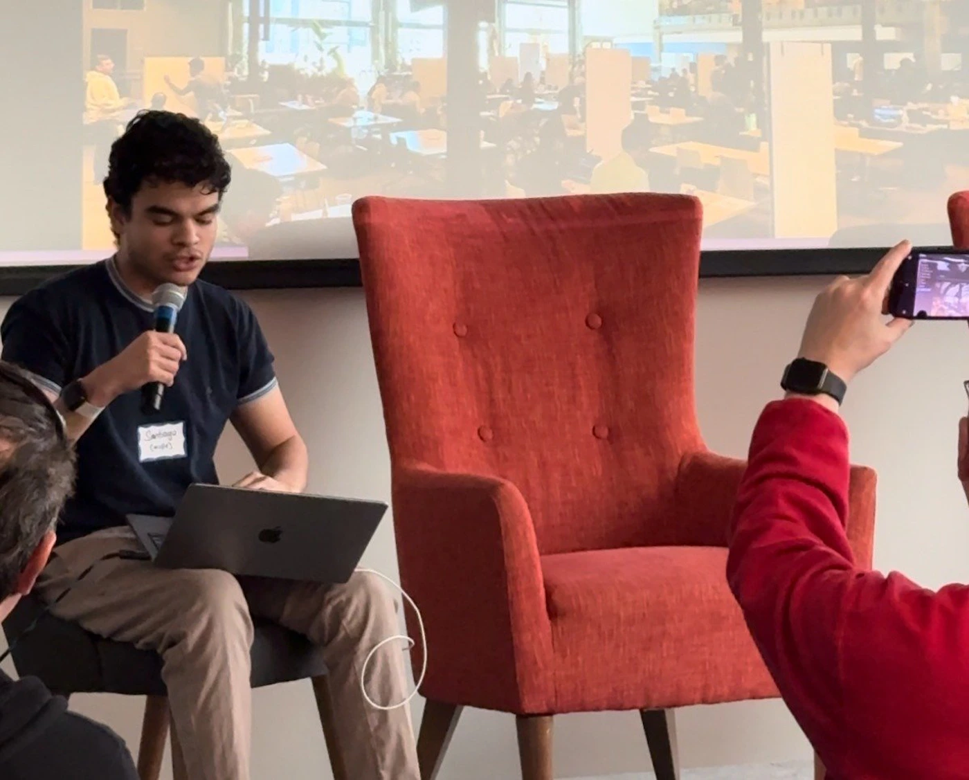 (Photo) A person sits in a chair, holding a microphone and a laptop, seemingly giving a presentation. Another person takes a picture of the speaker. Text: Santiago Torres person | indoor | presentation, audience, laptop, microphone | candid Note: The image is a real-life captured scene of a person giving a presentation, making it a photograph.