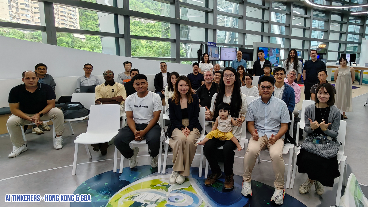 A group of diverse individuals, including a young child, are gathered for a photo in a modern, bright indoor space with large windows. Many are seated on white chairs, while others stand in the background, suggesting an event or meeting.. Text: AI TINKERERS - HONG KONG & GBA FOR SPONS