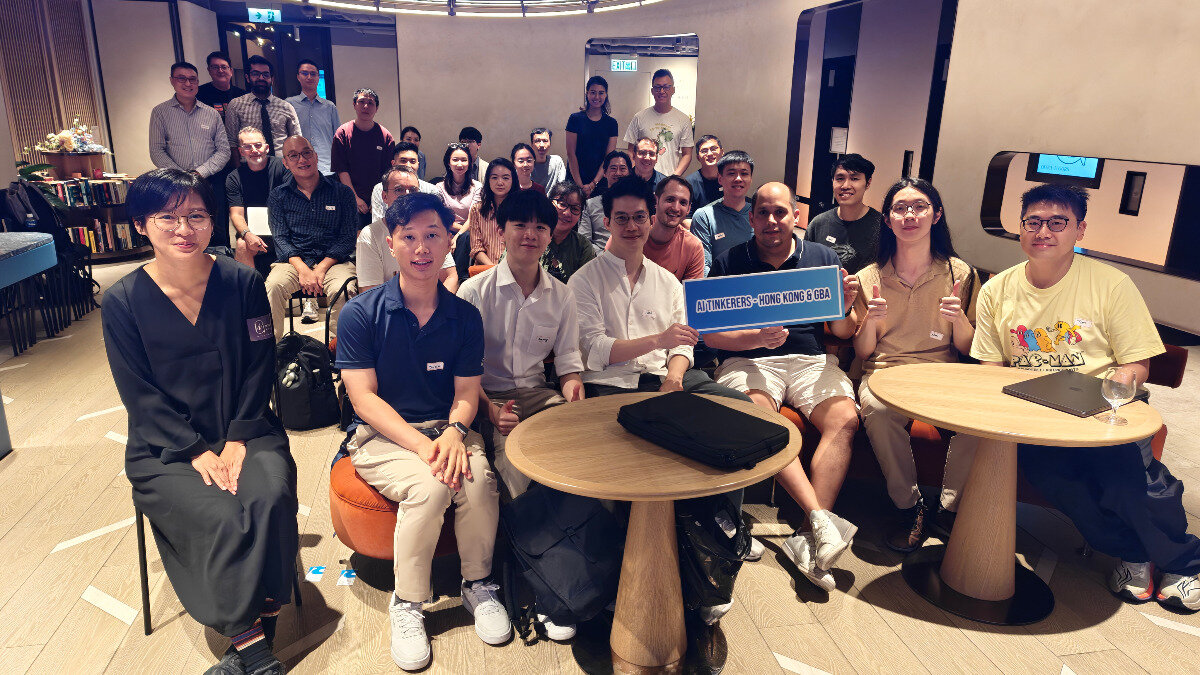 A large group of approximately thirty people, mostly young adults, are gathered indoors for a group photo in a modern lounge or event space. Several individuals in the front row are seated around small wooden tables, and one person holds a blue sign identifying the group.. Text: AI TINKERERS - HONG KONG & GBA PAC-MAN EXIT