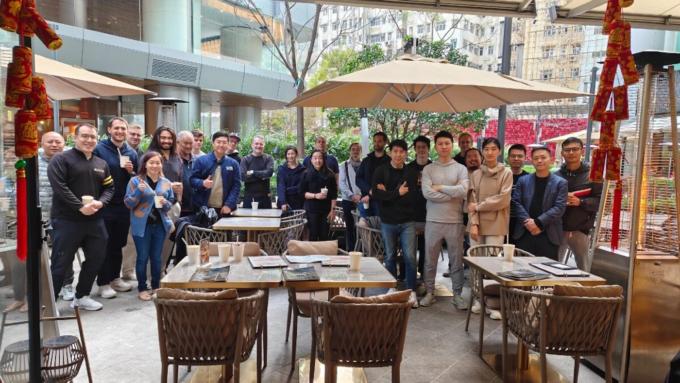 (Photo) A large group of approximately twenty people poses for a group photo in an outdoor, covered patio area of an urban restaurant, which is decorated with red Chinese New Year ornaments. Group of people, social gathering | Outdoor patio/cafe, urban environment | Large group (approx. 20), Chinese New Year decorations, outdoor seating area | Posed group shot | Colors: #A98C6D, #333333, #D0D0D0, #C83737 Note: The image is a photograph capturing a moment in time involving a large group of people in an outdoor urban setting.