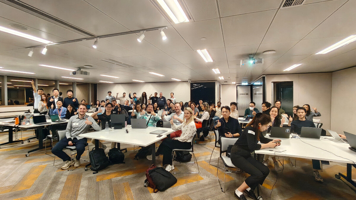 A large group of people are gathered in a modern office space, many of whom are seated at white tables with laptops and giving a thumbs-up gesture to the camera.. Text: CONFERENCE ROOM
FIRE EXIT
Build [unreadable] to the future together