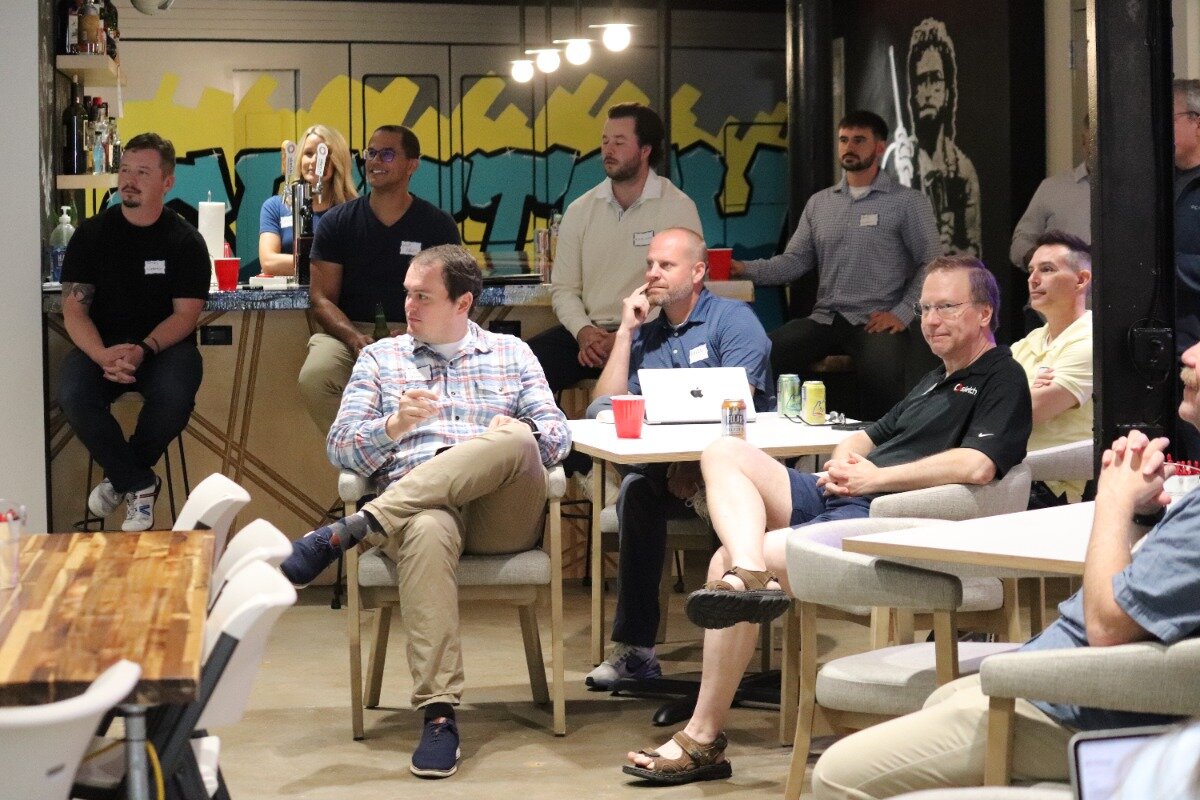 A group of people are gathered in a casual indoor setting, likely an office or event space, with some seated at tables and others standing near a bar area adorned with graffiti art.