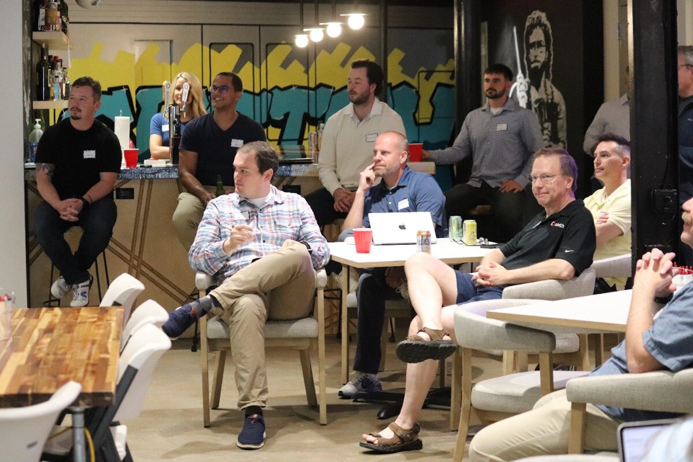 (Photo) A group of people are gathered in a casual indoor setting, likely an office or event space, with some seated at tables and others standing near a bar area adorned with graffiti art. group of people | indoor, casual event space or office | multiple people engaged in conversation or listening, some seated at tables with drinks and a laptop, others standing by a bar; a wall with yellow and teal graffiti art is visible in the background. | candid, event photography | Colors: #3B4E6C, #B09C77, #F5E040, #40B0B0, #E03030, #F0F0F0 Note: This image captures a real-world scene with multiple individuals and objects in a natural environment, characteristic of a photograph.