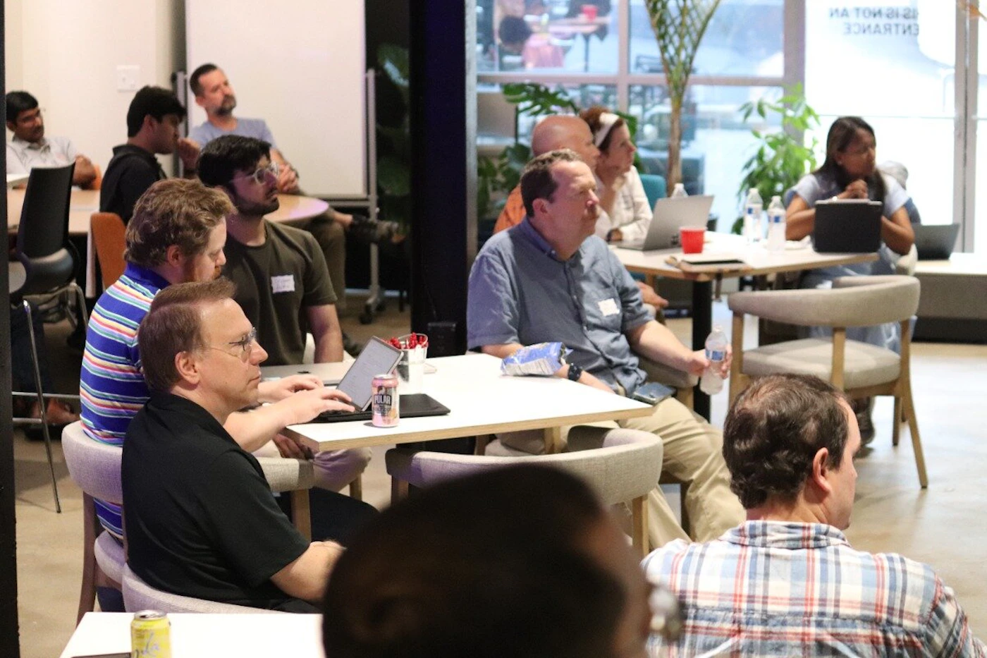 (Photo) A group of people are seated at tables in a modern, brightly lit indoor space, appearing to attend a meeting or presentation. Many are looking forward, and some have laptops or drinks on their tables. Text: AN EXIT IS NOT AN ENTRANCE, POLAR SELTZER group of people, indoor event/meeting | indoor, modern office or co-working space with large windows | multiple people, some with laptops, casual attire, bright lighting, plants | candid, documentary-style | Colors: #F8F8F8, #6A8FB7, #B0A080, #6B8E23 Note: The image is a direct capture of a real-world scene featuring multiple individuals engaged in an activity within an indoor environment, characteristic of a photograph.