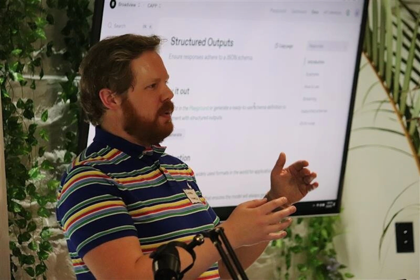 (Photo) The image shows a man with a beard, wearing a multi-colored striped polo shirt, standing and gesturing with his hands while giving a presentation. Behind him, a large screen displays text, including 'Structured Outputs' and 'Ensure responses adhere to a JSON schema'. Text: Bra[unreadable] CAPP: Search Structured Outputs Ensure responses adhere to a JSON schema Try it out in the Playground to generate a ready-to-use [unreadable] to [unreadable] with structured outputs tion overview quickstart examples pricing support changelog ADAM [unreadable] person presenting | indoor, likely a conference or event space | a man with a beard, wearing a striped polo shirt; a large screen displaying presentation slides; a microphone stand; and decorative green plants. | candid, documentary | Colors: #193478, #F0F0F0, #4A6E3B, #D94639 Note: The image is a direct capture of a real-world scene, featuring a person presenting in a room with a screen and plants, consistent with the characteristics of a photograph.
